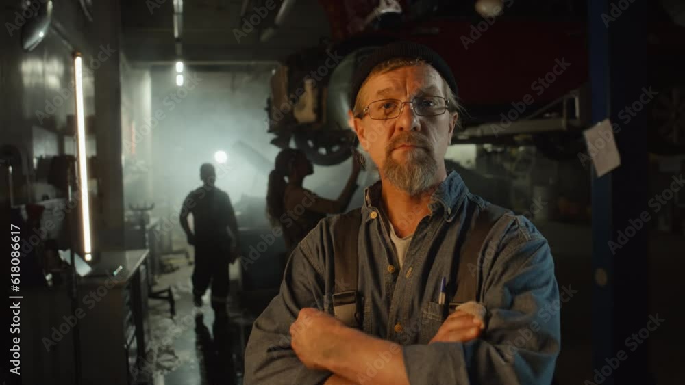 Tilt up portrait of mature Caucasian auto mechanic wiping his hands with piece of cloth then crossing his arms and looking at camera Tilt up portrait of mature Caucasian auto mechanic wiping his hands with piece of cloth then crossing his arms and looking at camera