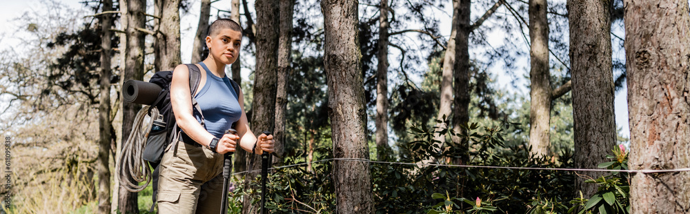 Short haired female tourist with backpack and travel equipment holding trekking poles and looking at camera while standing in summer forest, hiking for health and wellness concept, banner