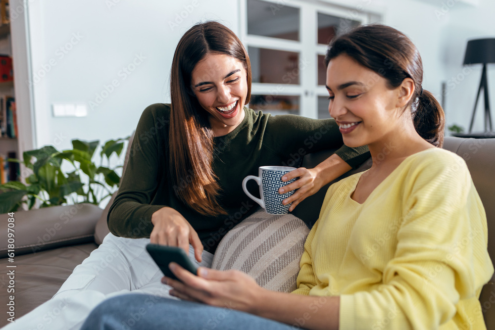 Two smiling young women talking while watching smartphone sitting on couch in the living room at home. Two smiling young women talking while watching smartphone sitting on couch in the living room at home.