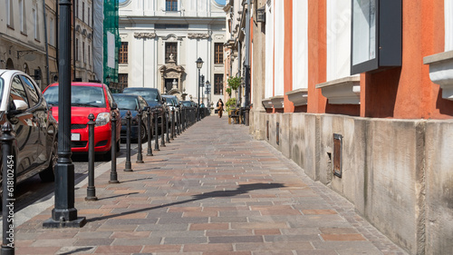 Many cars are parked in a queue on the side of an old European city street along a row of metal bollards, perspective of an old street with a female figure on the horizon, urban symmetry