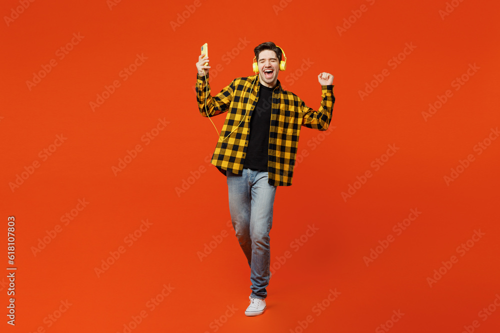 Full body young cheerful happy man wear yellow checkered shirt black t-shirt headphones listen to music use mobile cell phone isolated on plain red orange background studio portrait Lifestyle concept Full body young cheerful happy man wear yellow checkered shirt black t-shirt headphones listen to music use mobile cell phone isolated on plain red orange background studio portrait Lifestyle concept