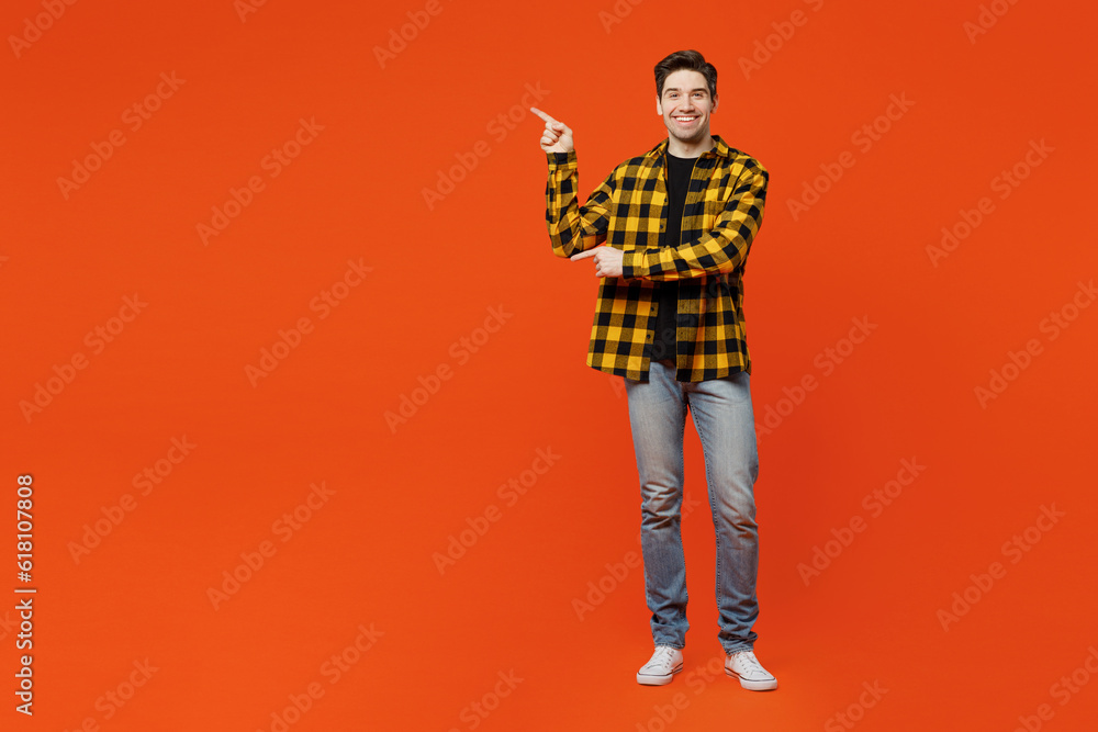 Full body young caucasian man wear yellow checkered shirt black t-shirt point index finger aside indicate on workspace area copy space mock up isolated on plain red orange background studio portrait.