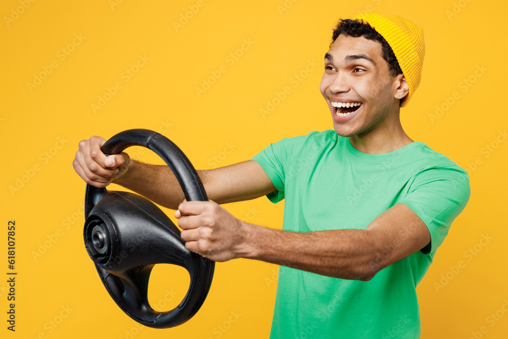 Side view young happy man of African American ethnicity wears casual clothes green t-shirt hat hold steering wheel driving car isolated on plain yellow background studio portrait. Lifestyle concept Side view young happy man of African American ethnicity wears casual clothes green t-shirt hat hold steering wheel driving car isolated on plain yellow background studio portrait. Lifestyle concept