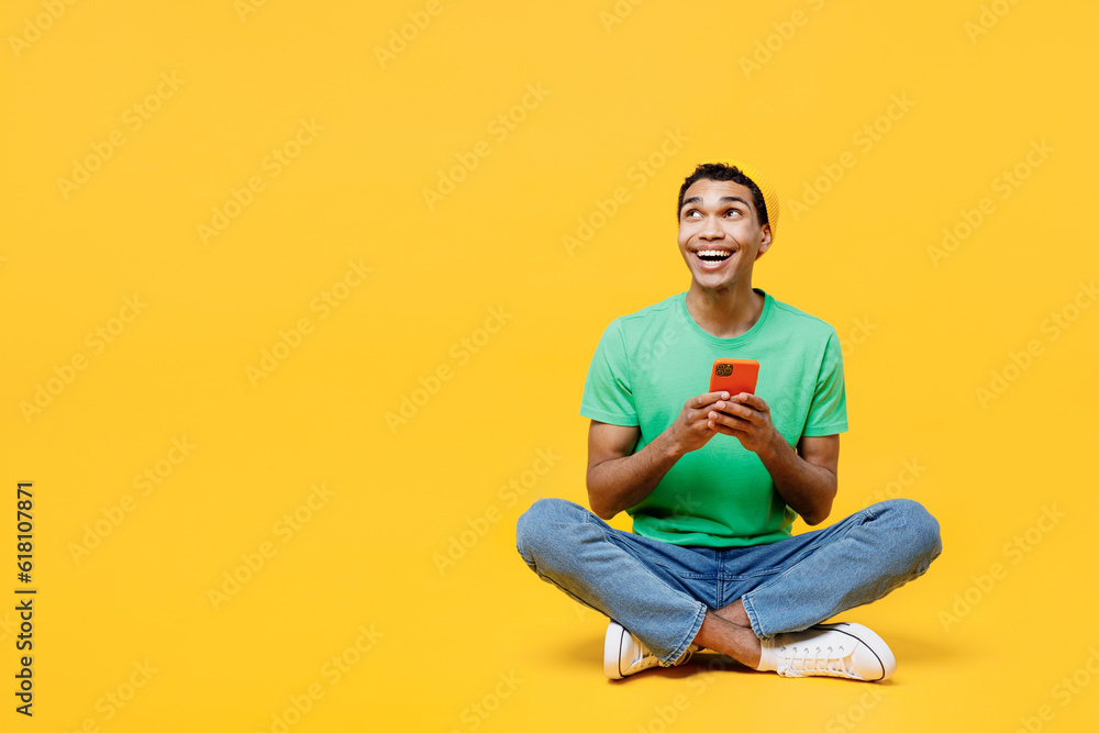 Full body young man of African American ethnicity he wears casual clothes green t-shirt hat sitting hold in hand use mobile cell phone look aside isolated on plain yellow background Lifestyle concept Full body young man of African American ethnicity he wears casual clothes green t-shirt hat sitting hold in hand use mobile cell phone look aside isolated on plain yellow background Lifestyle concept