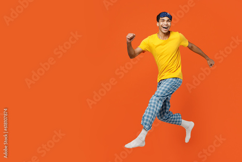 Side profile view young man wearing pyjamas jam sleep eye mask rest relax at home jump high running fast looking camera isolated on plain orange background studio portrait Good mood night nap concept