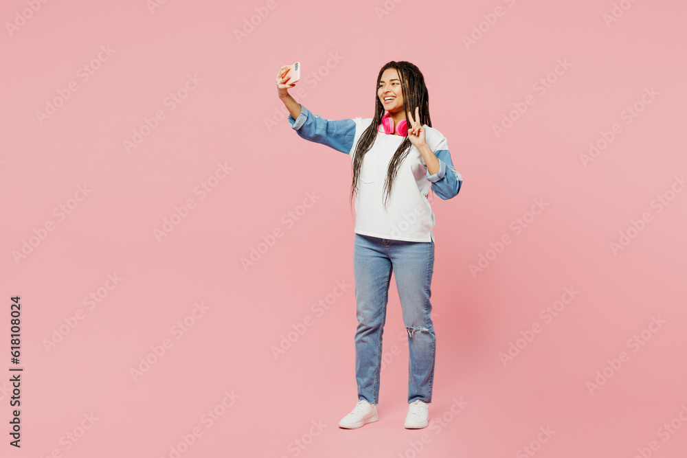 Full body young woman of African American ethnicity wear white sweatshirt casual clothes doing selfie shot on mobile cell phone show v-sign isolated on plain pastel pink background. Lifestyle concept. Full body young woman of African American ethnicity wear white sweatshirt casual clothes doing selfie shot on mobile cell phone show v-sign isolated on plain pastel pink background. Lifestyle concept.