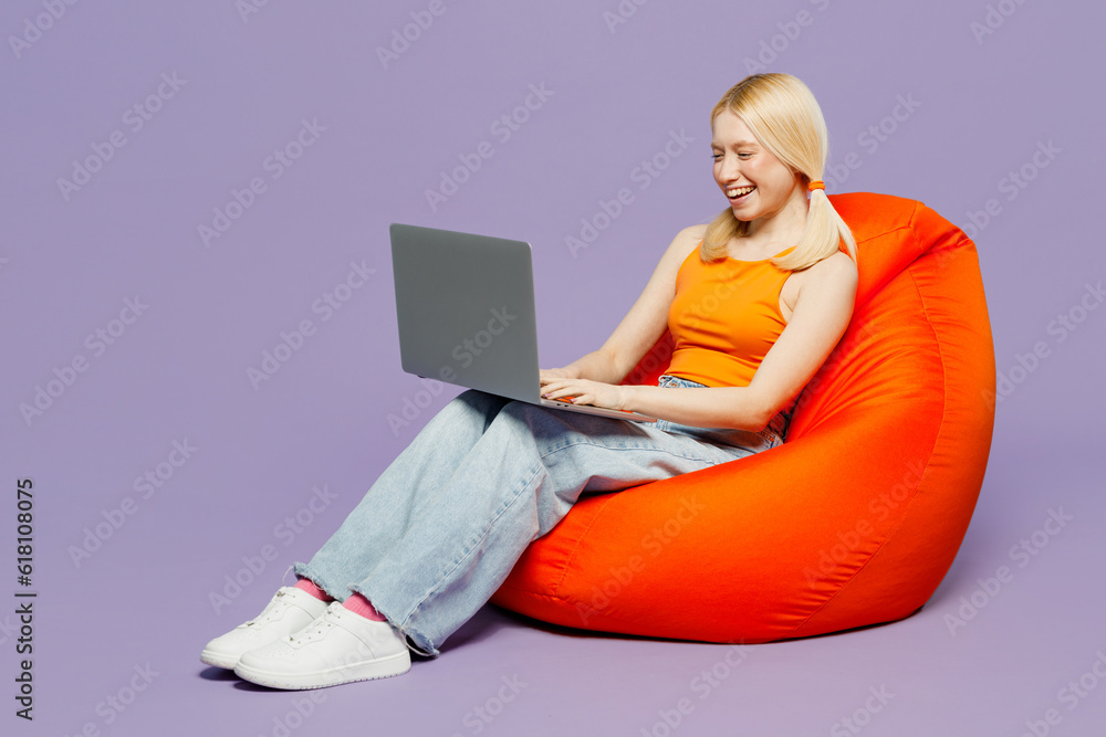 Naklejka premium Full body young blonde IT woman wear orange tank shirt casual clothes sit in bag chair hold use work on laptop pc computer isolated on plain pastel light purple background studio Lifestyle concept.