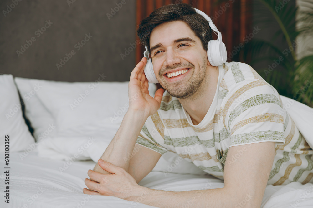 Young smiling happy man wear casual clothes t-shirt pajama headphones listen to music lying in bed rest relax spend time in bedroom home in own room hotel wake up dream be in reverie good mood day. Young smiling happy man wear casual clothes t-shirt pajama headphones listen to music lying in bed rest relax spend time in bedroom home in own room hotel wake up dream be in reverie good mood day.