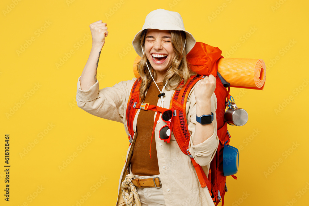 Young woman carry backpack with stuff mat doing winner gesture clench fist isolated on plain yellow background. Tourist leads active lifestyle walk on spare time. Hiking trek rest travel trip concept