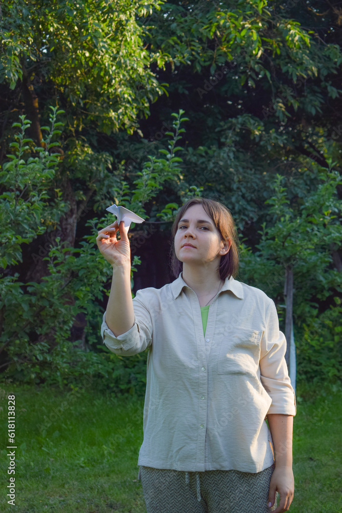 Balance concept. Front view happy adult woman launches paper airplane in bright sun rays in beautiful landscape garden at sunset. Serene female person uses origami to express harmony state Balance concept. Front view happy adult woman launches paper airplane in bright sun rays in beautiful landscape garden at sunset. Serene female person uses origami to express harmony state