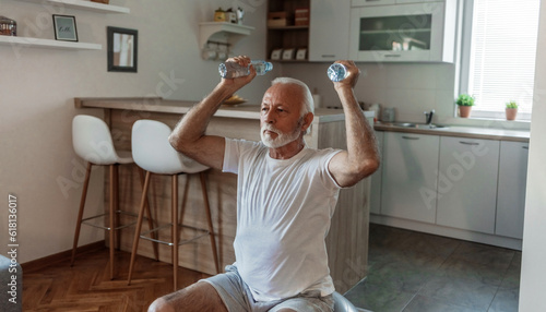 Bearded senior man doing exercises with water bottles sitting on fitball stretching at home indoor.