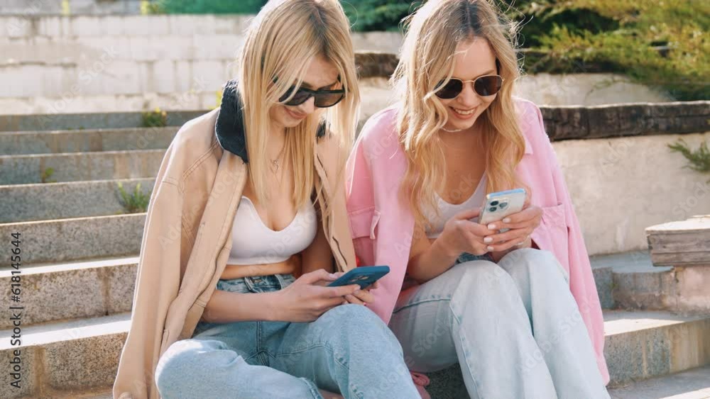 Two young beautiful smiling hipster female. Carefree women sitting at the stairs in the street in sunglasses. Models having fun. They looking at cellphone screen, using mobile apps, hold smartphone Two young beautiful smiling hipster female. Carefree women sitting at the stairs in the street in sunglasses. Models having fun. They looking at cellphone screen, using mobile apps, hold smartphone