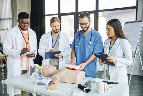 multiethnic students writing next to instructor showing wound care simulator near CPR manikin, automated defibrillator and medical equipment in training room, emergency situations response concept