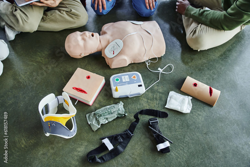 cropped view of multiethnic participants of first aid seminar near CPR manikin, defibrillator, wound care simulators, bandages, compression tourniquets and neck brace in training room, top view