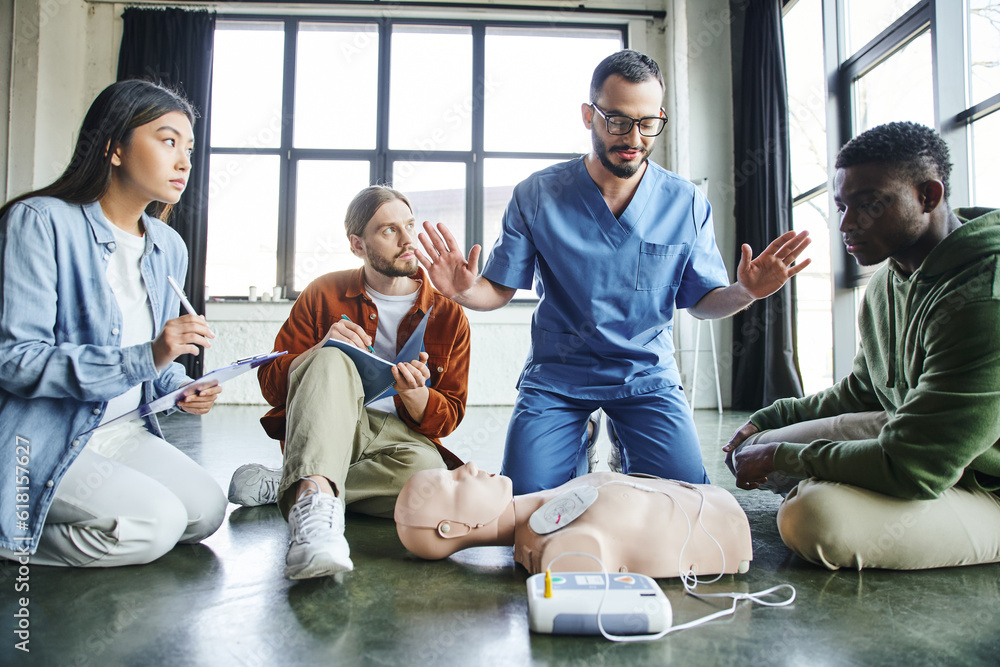 professional paramedic gesturing and talking to multiethnic participants near CPR manikin and defibrillator during first aid seminar in training room, health care and life-saving techniques concept - obrazy, fototapety, plakaty 