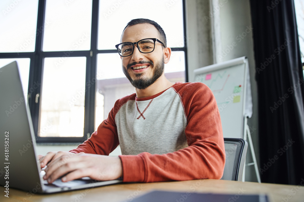 Fototapeta premium youthful and smiling bearded businessman in eyeglasses typing on laptop and looking at camera in front of flip chart with graphs on blurred background in contemporary office