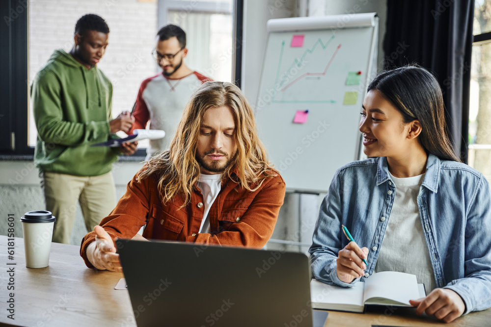 Fototapeta premium long haired man pointing at laptop near smiling asian woman while interracial colleagues working near flip chart with graphs on blurred background, multicultural collaboration in modern office