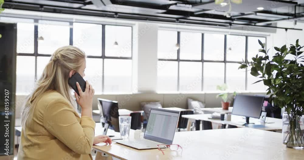 Happy plus size caucasian casual businesswoman using laptop and talking on smartphone at desk Happy plus size caucasian casual businesswoman using laptop and talking on smartphone at desk