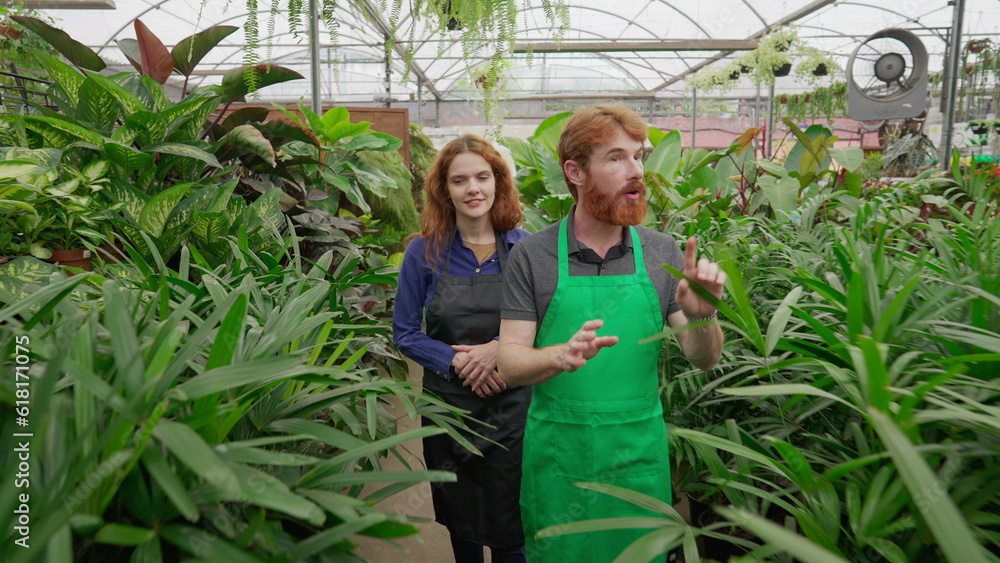 Obraz premium Two employees walking through isle inside plant store. man and woman staff wearing aprons, male employee explaining Flower Shop business