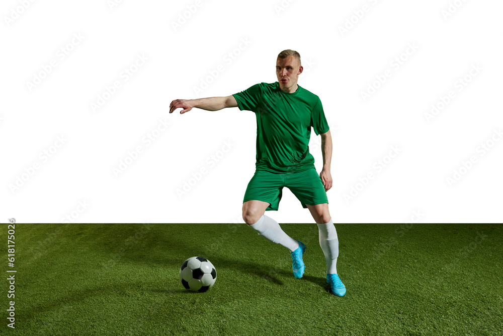 Concentrated young man, football player in green uniform dribbling ball, training against white background. Concept of professional sport, action, lifestyle, competition, hobby, training, ad