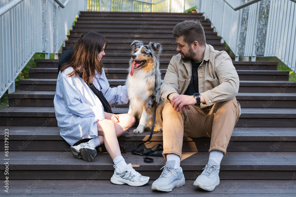 Obraz premium Happy young woman and man sitting with their aussie dog outdoors in urban park. Couple and their pet australian shepherd