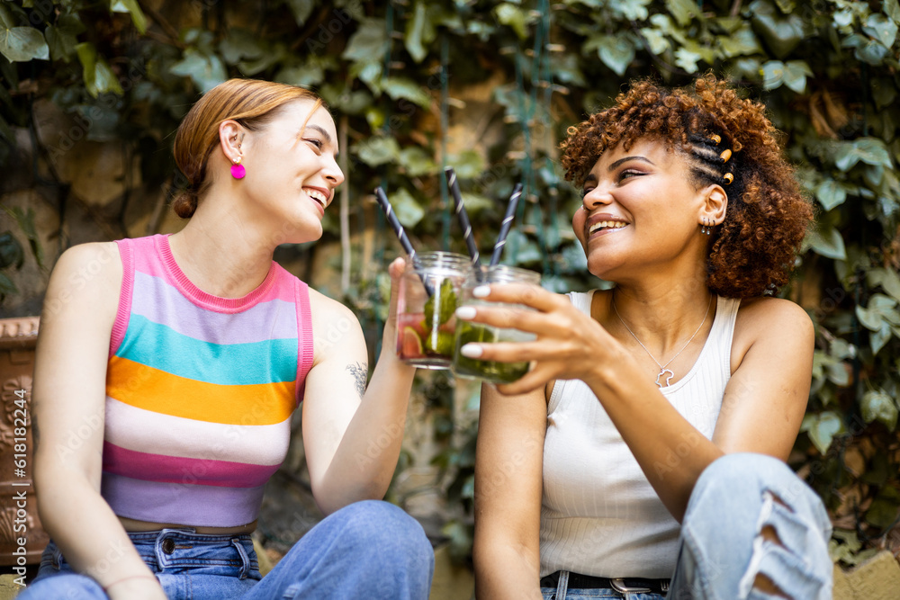 Two cute multiracial girls are happily toasting with some flavored cocktails in a place with vegetation. Concept of multiethnic couple drinking mojitos, toast with combined. - obrazy, fototapety, plakaty 