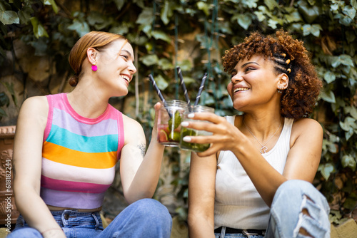Two cute multiracial girls are happily toasting with some flavored cocktails in a place with vegetation. Concept of multiethnic couple drinking mojitos, toast with combined.