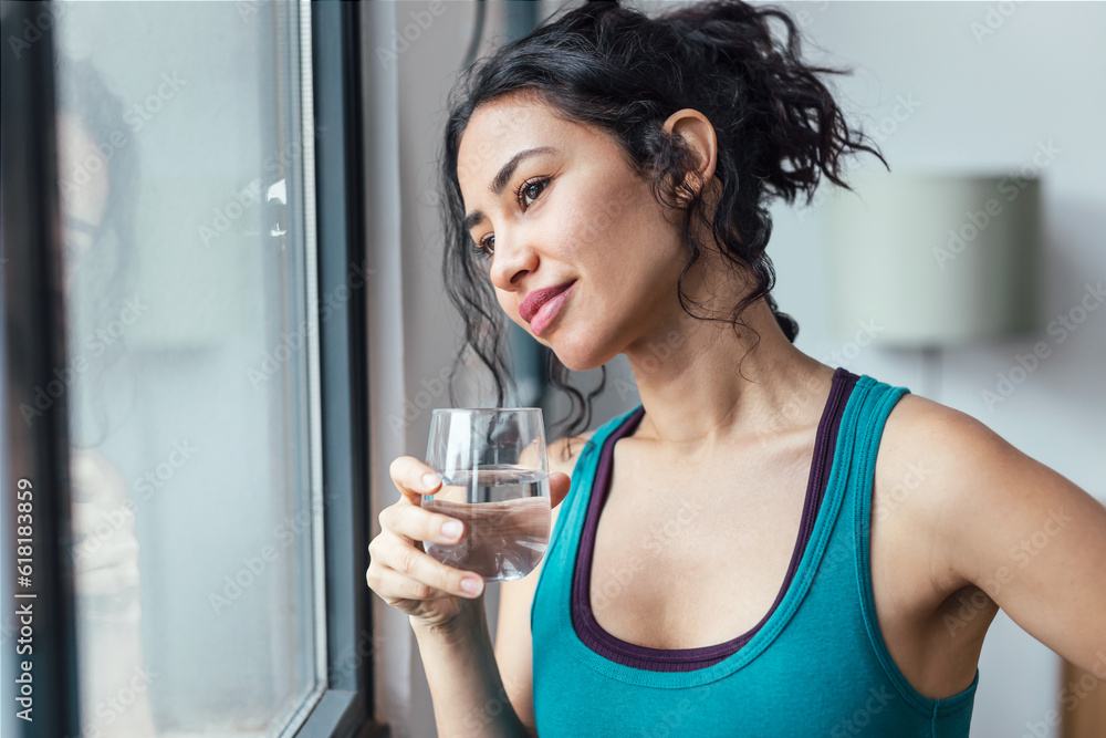 Φωτογραφία Beautiful sporty woman drinking a glass of water while standing near the window