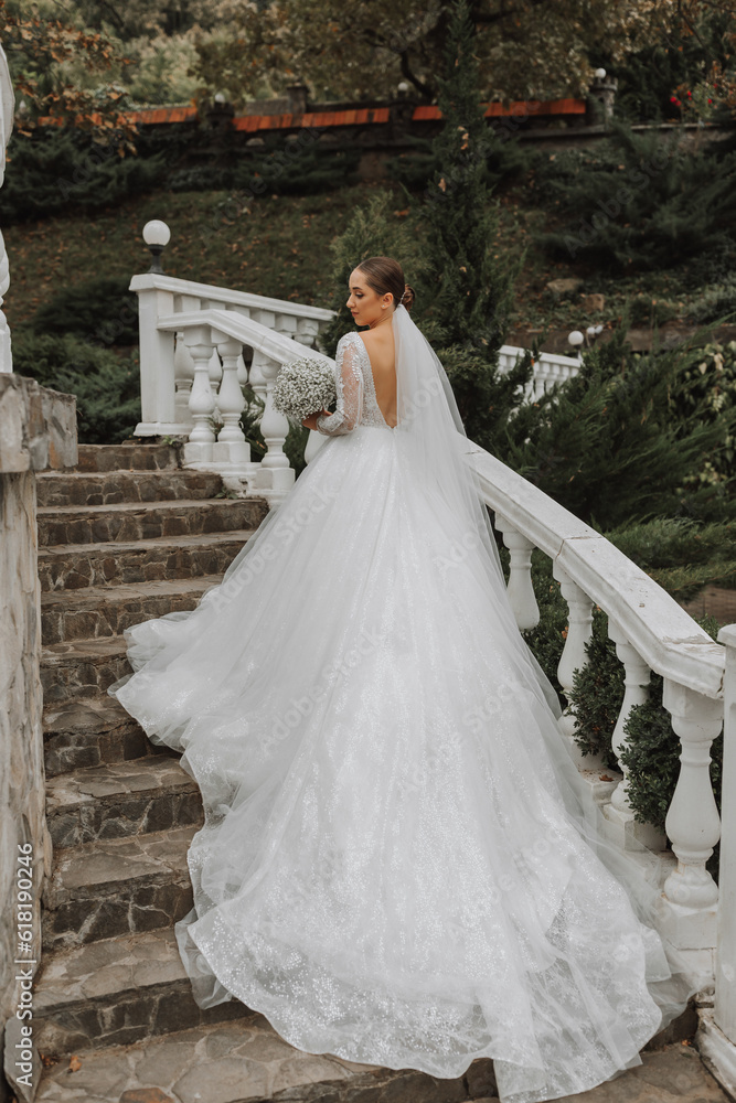 Chic image of the bride, a wedding dress with a long train. The bride with a bouquet of flowers climbs up the steps in the garden. Open shoulders Chic image of the bride, a wedding dress with a long train. The bride with a bouquet of flowers climbs up the steps in the garden. Open shoulders