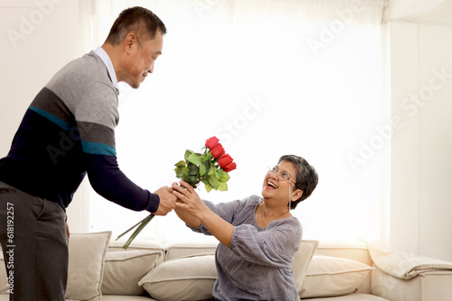 Romantic elderly man gives rose flower bouquet to his wife while sitting in living room, happy smiling Asian senior mature retired couple celebrate Anniversary together at home, retirement lifestyle.