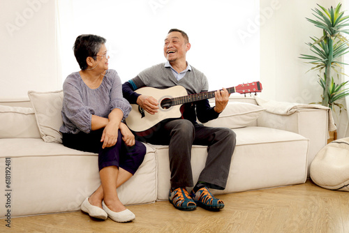 Happy smiling Asian elderly couple relaxing and playing guitar while sitting on sofa in the living room, romantic senior mature retired couple having fun together at home, retirement lifestyle concept