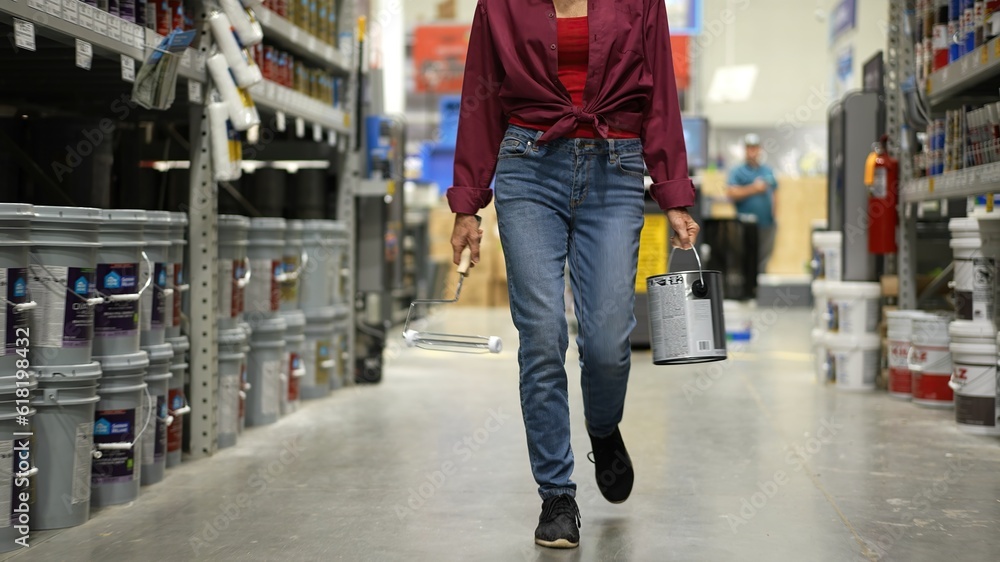 Obraz premium Woman shown from waist down, carrying can of paint and roller walking down paint aisle in hardware store