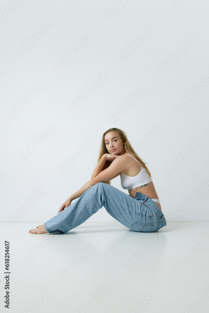 Tenderness. Beautiful young blonde woman with slim body posing in white top and jeans against grey studio background. Concept of beauty, body and skin care, health, fitness, wellness, ad