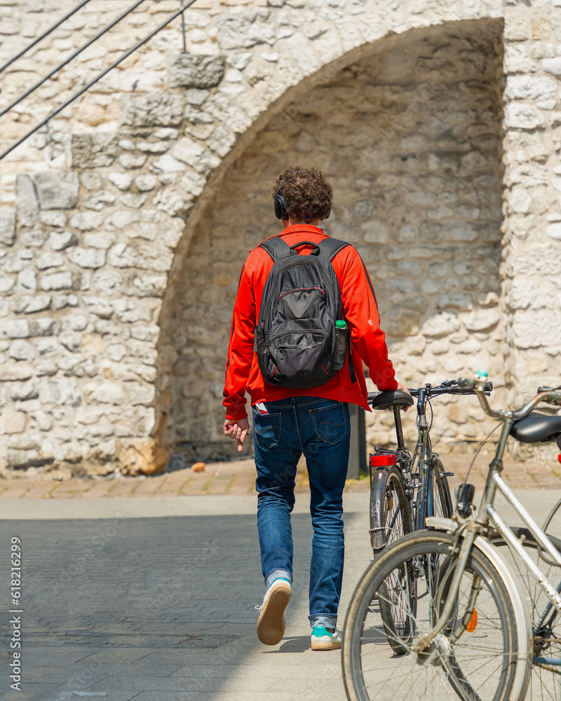A young man in a red jacket walks along an old European street with a bicycle and an electronic cigarette in his hand against the background of a stone medieval wall, healthy lifestyle and bad habits