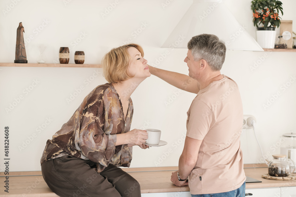 Fototapeta premium middle age woman reaching out to kiss her husband while sitting on the kitchen table and drinking tea, happy mature family, tender and affectionate couple.