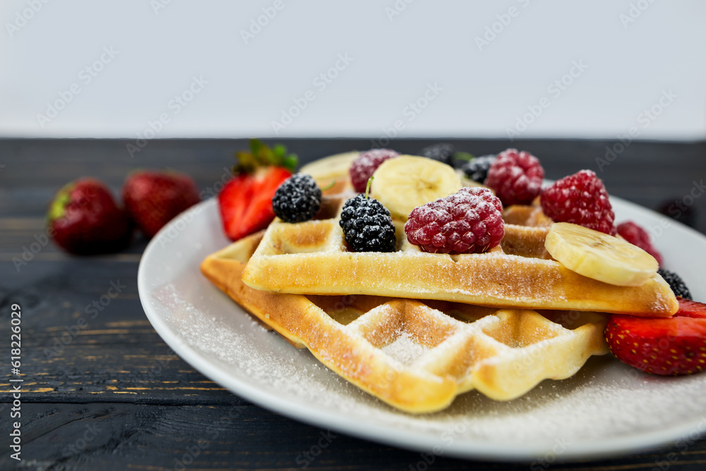 Belgian waffles with summer berries and powdered sugar in a white plate on a dark wooden background. Belgian waffles with summer berries and powdered sugar in a white plate on a dark wooden background.