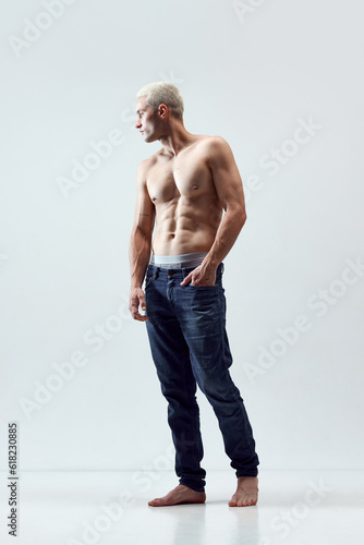 Full-length portrait of handsome young man with blonde hair, musuclar body posing shirtless in jeans against grey studio background. Concept of male natural beauty, body care, health, sport, ad photo