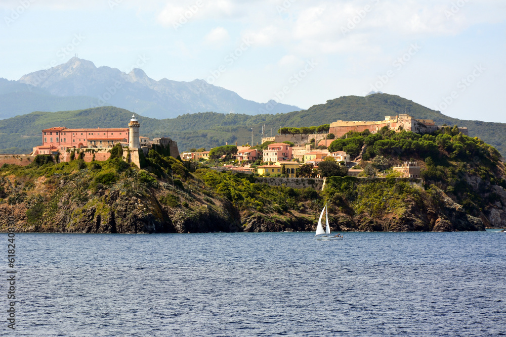 The lighthouse and buildings of the old fort are located on the picturesque Mediterranean seashore. Mountains are behind them in the distance