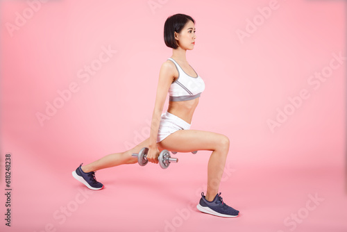 Sporty woman holding two dumbells and doing leg lunges for leg muscle training isolated on pink background. Woman with a healthy lifestyle.