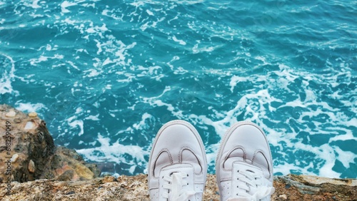 Close-up feet of resting tourist in white shoes on rocky seashore on nature. Relaxing feet outdoors after walk. Clean sneakers with laces in sunlight. Recovery of strength. Leg fatigue. Close up.