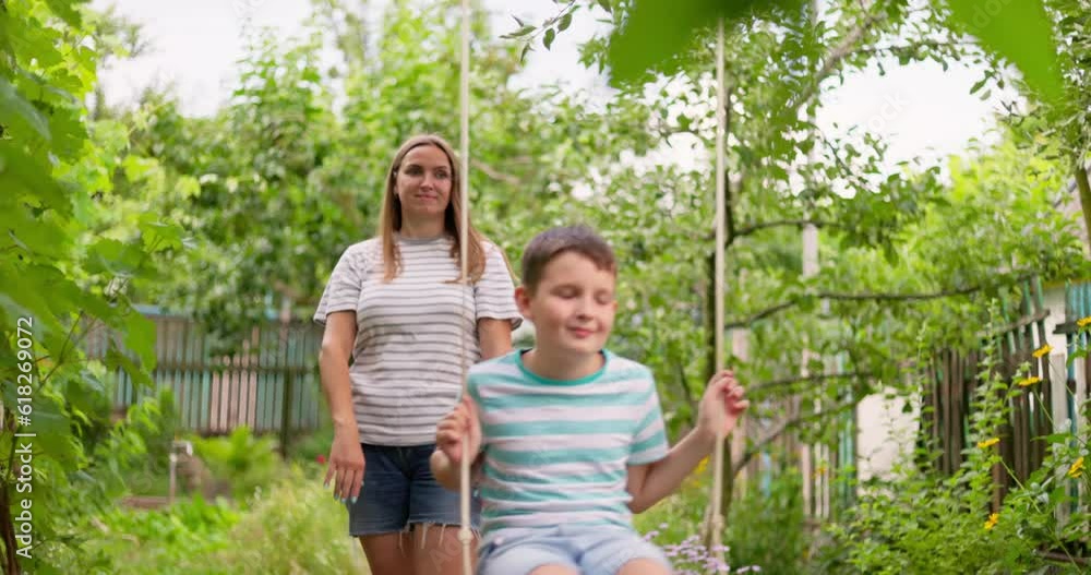 Happy family, mother and child play on wooden swing in garden under tree Happy family, mother and child play on wooden swing in garden under tree