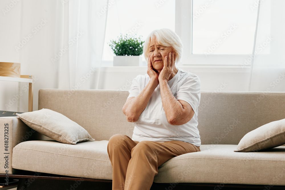 Elderly woman severe pain in the head sitting on the couch, health problems in old age, poor quality of life. Grandmother with gray hair holding her head, migraine and high blood pressure. Elderly woman severe pain in the head sitting on the couch, health problems in old age, poor quality of life. Grandmother with gray hair holding her head, migraine and high blood pressure.