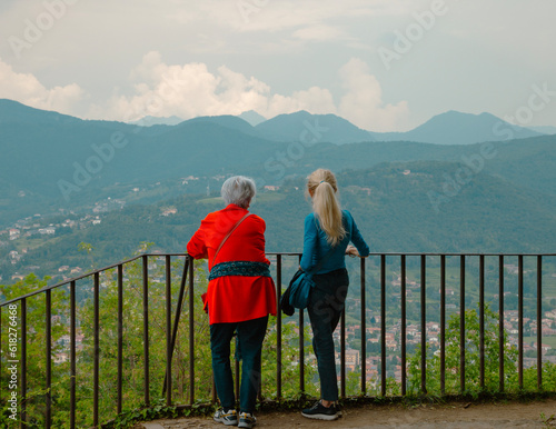 Rear view of two stylish senior women enjoying a beautiful landscape in italian city of Bergamo,  sitting near a fence and facing scenic mountains and green fields