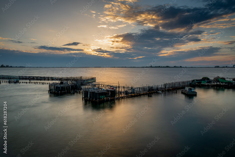 Long exposure of a fish farm during sunset with the sun lighting up the clouds and casting light on the water.