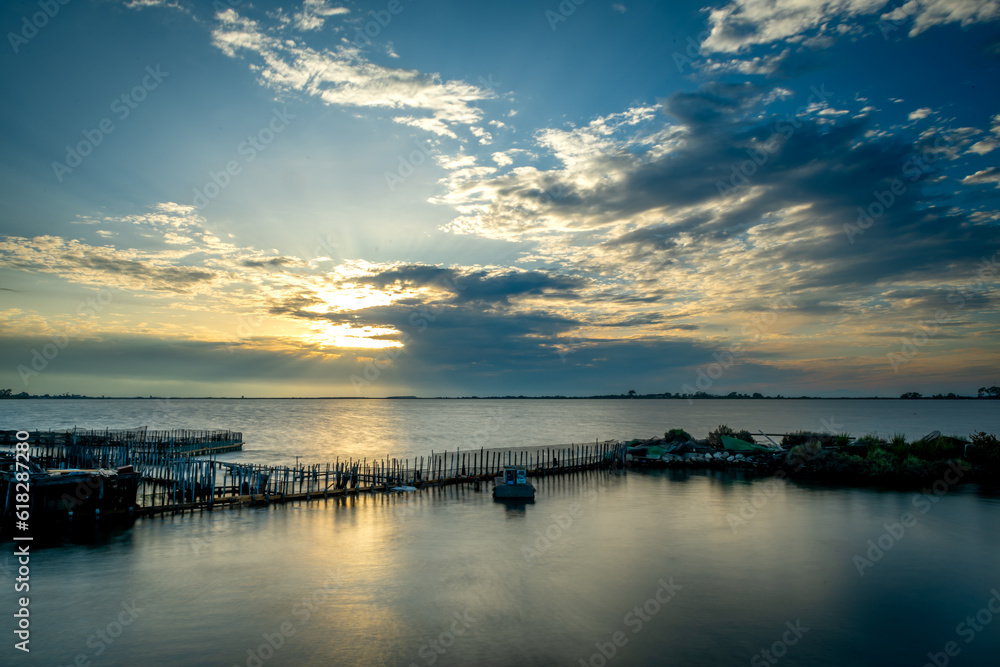 Long exposure of a fish farm during sunset with the sun lighting up the clouds and casting light on the water.