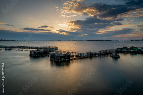 Long exposure of a fish farm during sunset with the sun lighting up the clouds and casting light on the water. © Ming