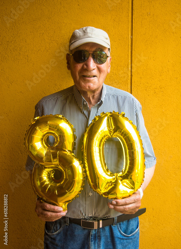 Old man holding a 80's birthday numbers balloons smiling and celebrating on his party. Wearing a cap, sunglasses, shirt and jean. Yellow wall on background