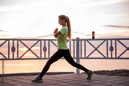 Young woman doing lunges, squats on warm-up in morning at sunrise by sea. Sportswoman in sportswear