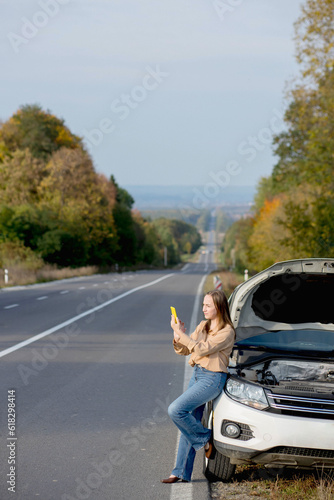 Young woman driver standing near a broken car with popped up hood and call on the poor mobile network coverage in the autumn