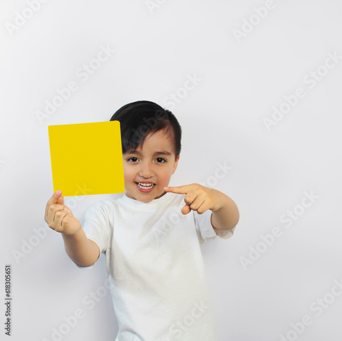 A beautiful latin boy with big smile holds a yellow piece of paper in a white t-shirt on a white background, points at it with his finger. Space for text, banners, label surfaces, and ads. 